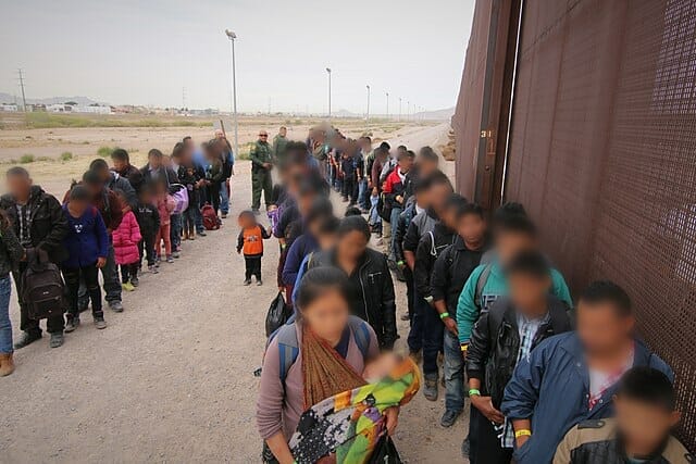 Nota sobre la nueva propuesta del gobierno para mejorar el sistema de asilo en Estados Unidos. La foto es de una fila de inmigrantes junto al muro fronterizo con México.